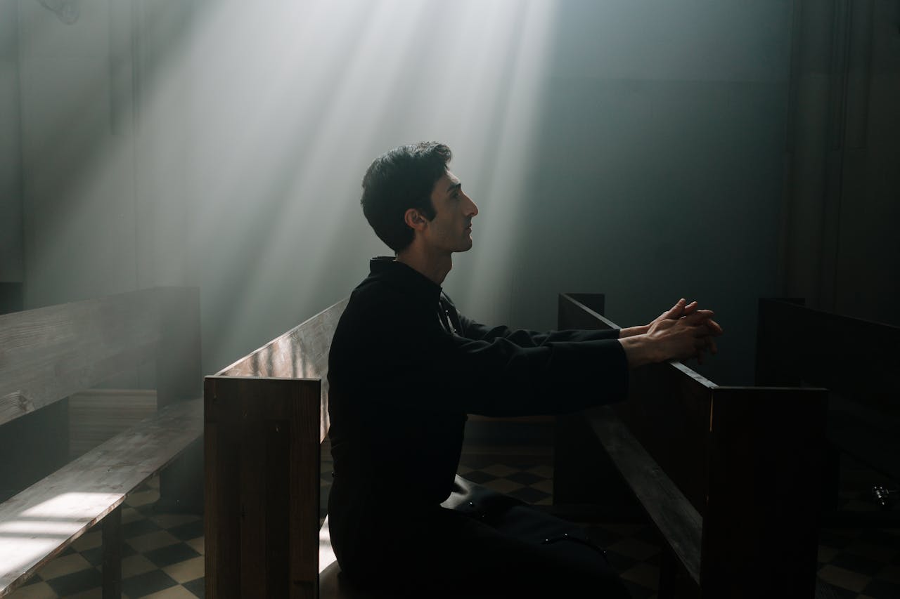 A man sits praying in sunlight inside a dramatic church setting.